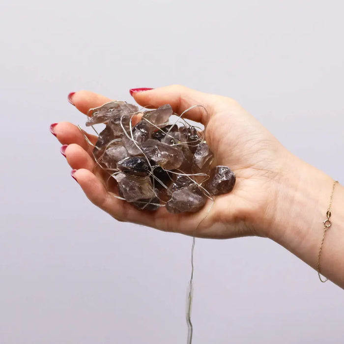 A hand holds smoky quartz crystals wrapped in silver wire as part of gemstone enchantment lights with citrine