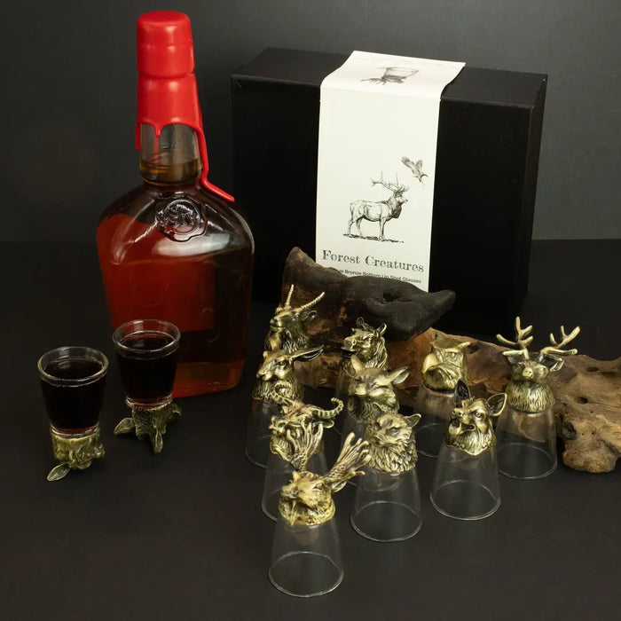 Set of clear shot glasses with intricate antique bronze mystical creature stoppers next to red-lidded bottle and gift box