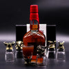 A tall amber liquor bottle with a red cap and gold neck detailing next to six antique bronze mystical creatures shot glasses