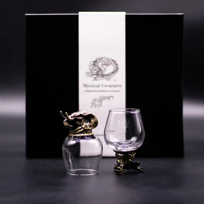 A clear glass bottle with a golden dragon stopper and base next to a crystal snifter on an antique bronze stand with mystical creatures