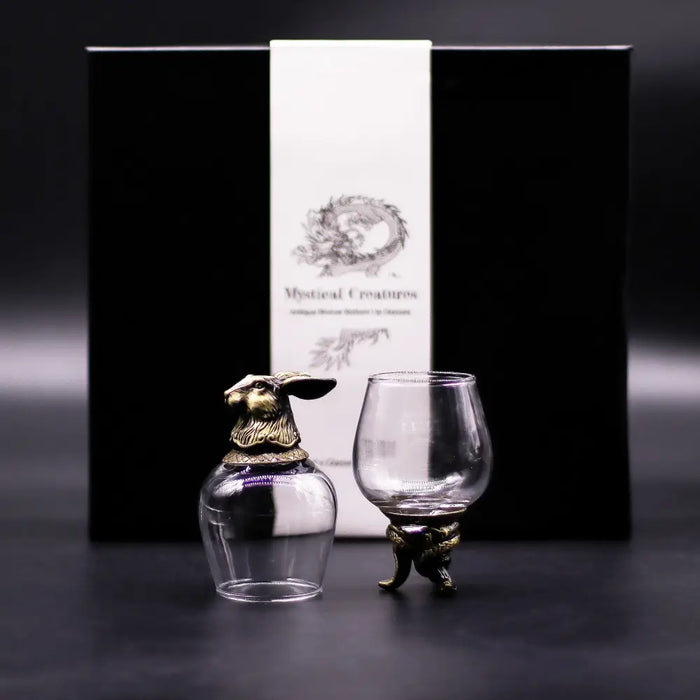 A glass decanter with a golden rabbit stopper and matching footed glass featuring antique bronze mystical creatures details