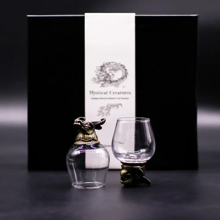 A clear glass decanter with an antique bronze golden dragon stopper and base next to an empty crystal tasting glass