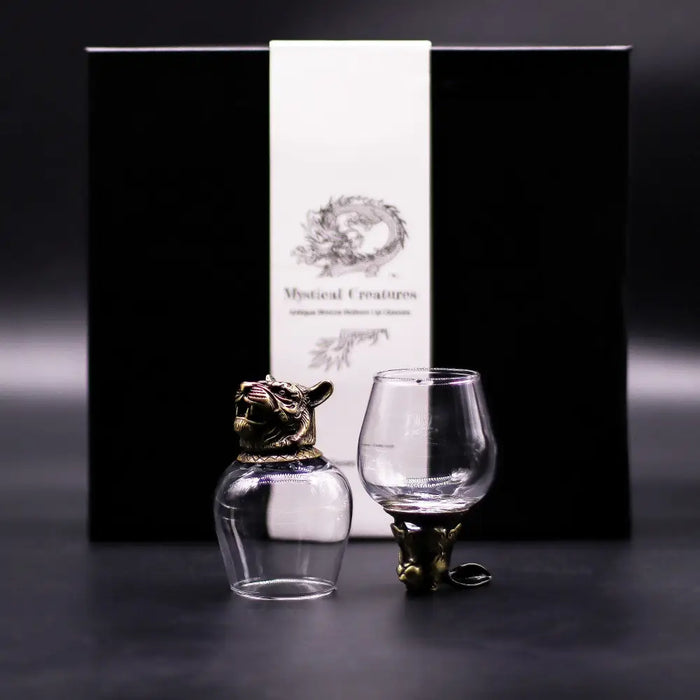 A clear glass bottle with antique bronze tiger-head stopper and a matching mystical creatures stemmed glass foot