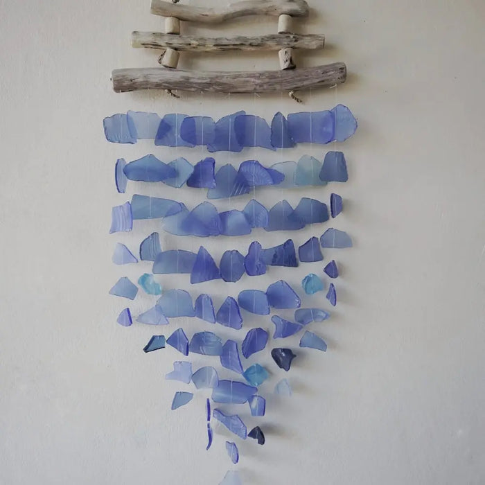 A driftwood wind chime with three sticks made of layered blue sea glass pieces hanging from rustic wood