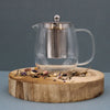 Glass infuser teapot with clear glass body and stainless steel infuser and lid knob resting on wooden slice