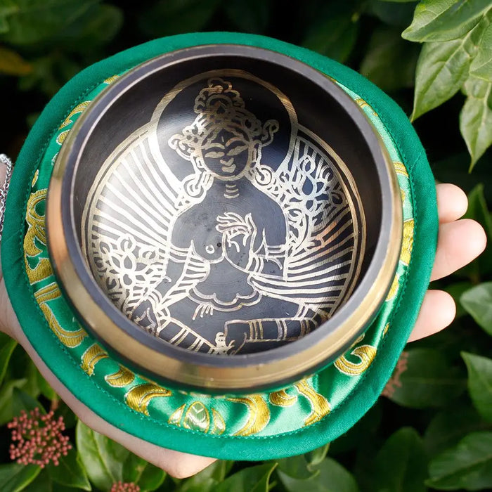 A hand holds a green tara singing bowl with a green silk lining and silver gilded deity design inside a dark bowl