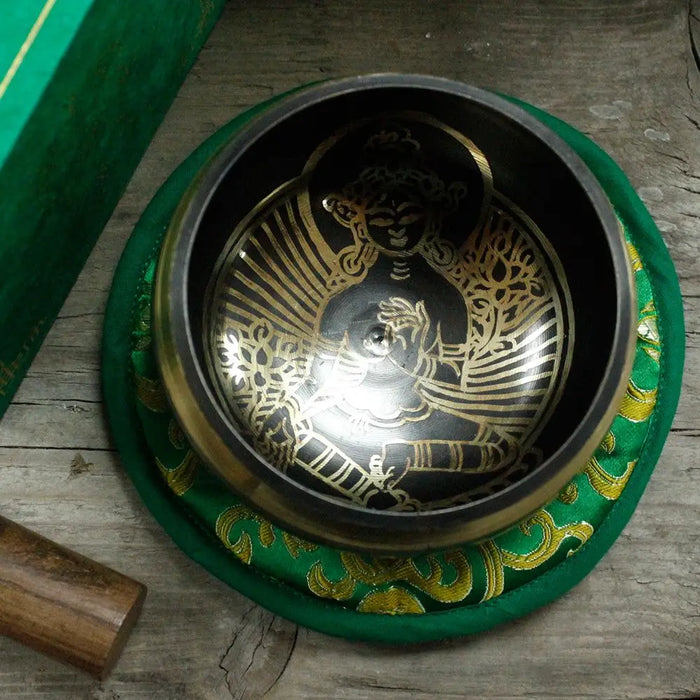 A black and gold tibetan singing bowl with green tara engravings on a green embroidered cushion 10cm