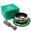 Black and gold green tara singing bowl with intricate engravings on a green embroidered mat beside its branded box