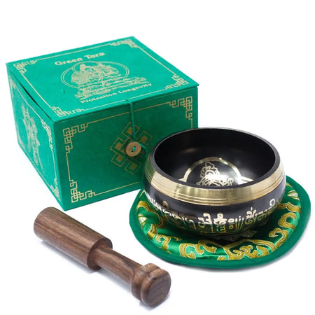 Black and gold green tara singing bowl with intricate engravings on a green embroidered mat beside its branded box