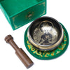 A black and gold green tara singing bowl with engraved buddha design resting on a green embroidered mat