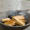 A smoldering palo santo stick burning in a dark bowl with light brown holy wood and white sage sticks nearby