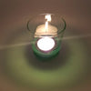 A clear glass votive tealight holder with a green base holding a lit white tea light candle