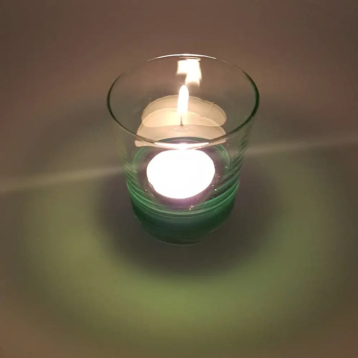 A clear glass votive tealight holder with a green base holding a lit white tea light candle