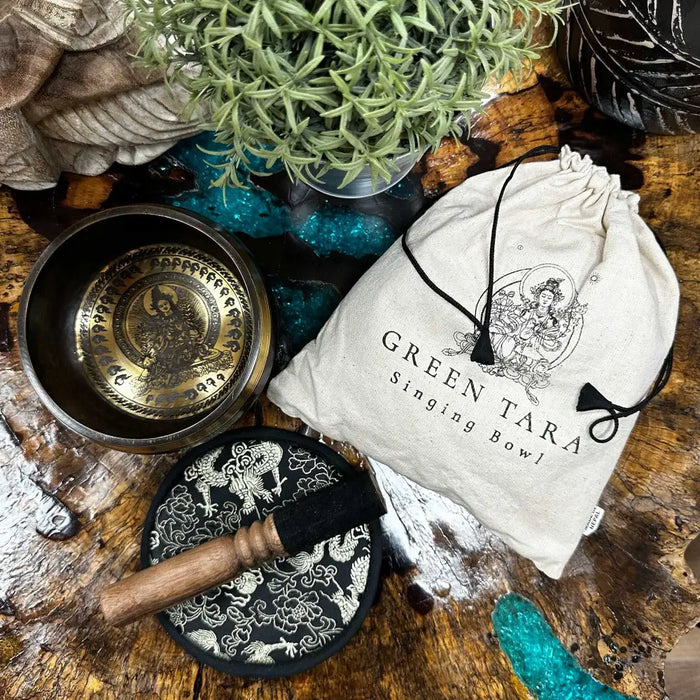 A green tara engraved singing bowl in brass with intricate golden designs and a black and white patterned mallet beside it