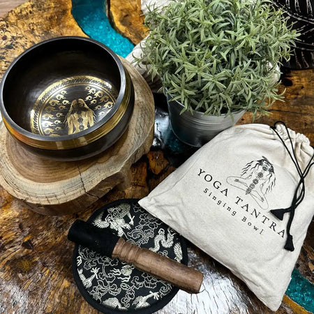A dark bronze hand-beaten engraved singing bowl with gold figure engravings resting on a wooden stand in a bowl gift set
