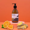 An amber glass bottle of ancient wisdom body lotion with ginger and orange scent on a pink pedestal with black pump