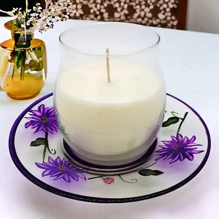 A creamy white scented candle in a clear glass jar on a hand-painted glass candle plate with purple wild flowers