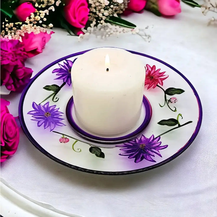 A white cylindrical candle with a lit wick on a hand painted glass candle plate with purple wild flowers design