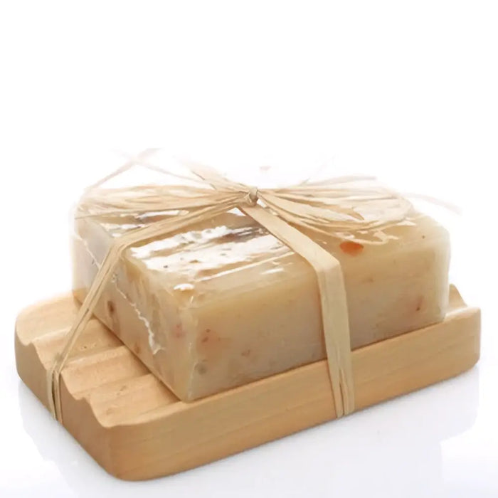 A rectangular off-white soap bar with natural specks wrapped in plastic and tied with twine on a hemu wood soap dish