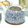 A white ceramic teapot with blue floral patterns brown glazed rims and a woven bamboo handle for herbal tea