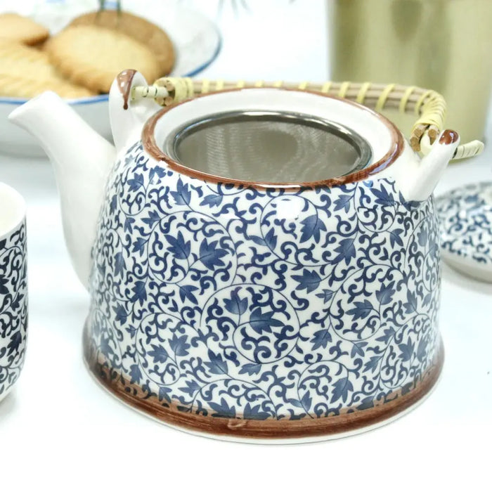 A white ceramic teapot with blue floral patterns brown glazed rims and a woven bamboo handle for herbal tea