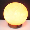 A warm glowing himalayan salt lamp ball with a smooth translucent surface resting on a dark wooden base highlighting its hygroscopic properties