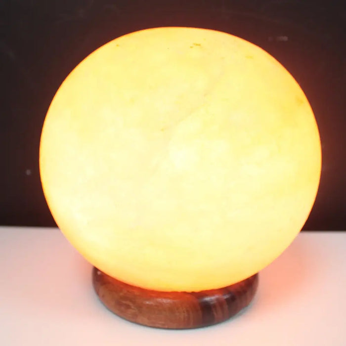 A warm glowing himalayan salt lamp ball with a smooth translucent surface resting on a dark wooden base highlighting its hygroscopic properties