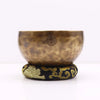A hand-forged brass tibetan singing bowl with hammered texture and symbol on a black and gold embroidered hoop cushion