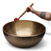 A hand holds a wooden mallet with a red head above a large hammered bronze feet standing bowl 50cm
