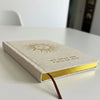 A cream-colored linen-bound ivory gratitude journal with gold sun emblem and yellow interior with ribbon bookmark