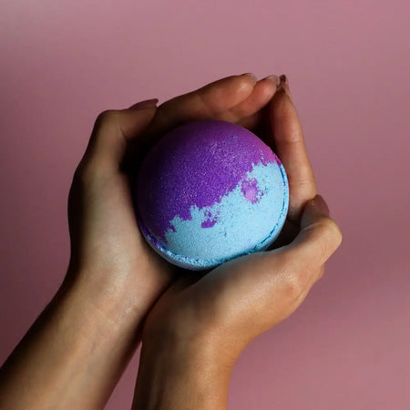 A jumbo bath bomb in vibrant purple and blue with a glittery texture held in hands containing sodium lauryl sulfoacetate