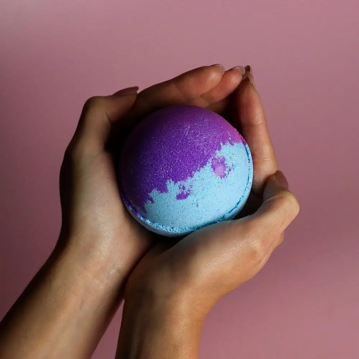 A jumbo bath bomb in vibrant purple and blue with a glittery texture held in hands containing sodium lauryl sulfoacetate