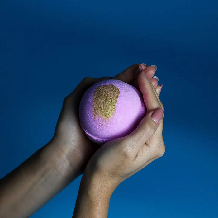 A jumbo bath bomb pink charlotte in soft lavender with gold foil held in hands featuring sodium lauryl sulfoacetate and peel oil