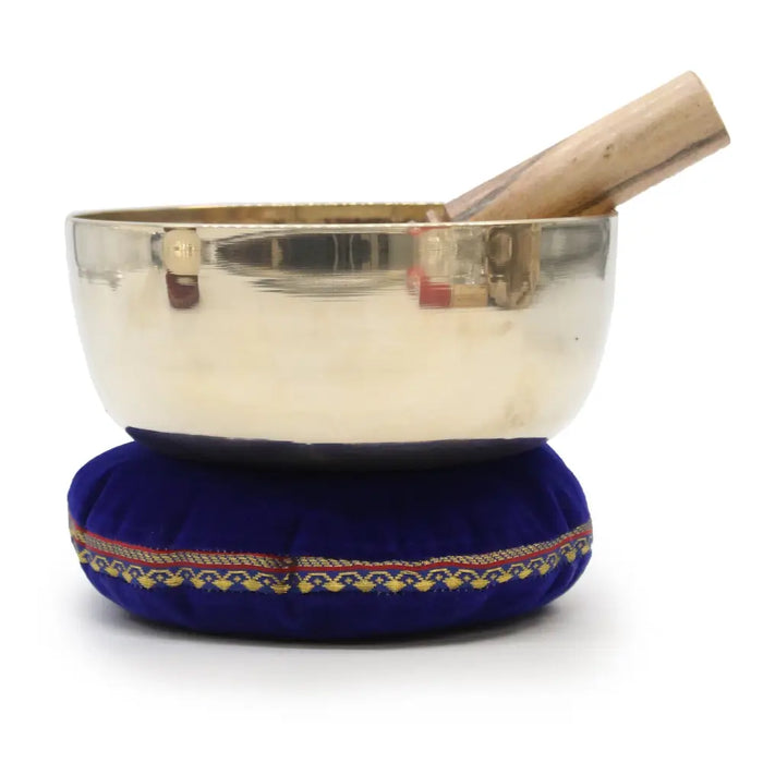 Large brass singing bowl with a shiny silver finish and wooden mallet resting on a plush royal blue cushion singing bowls