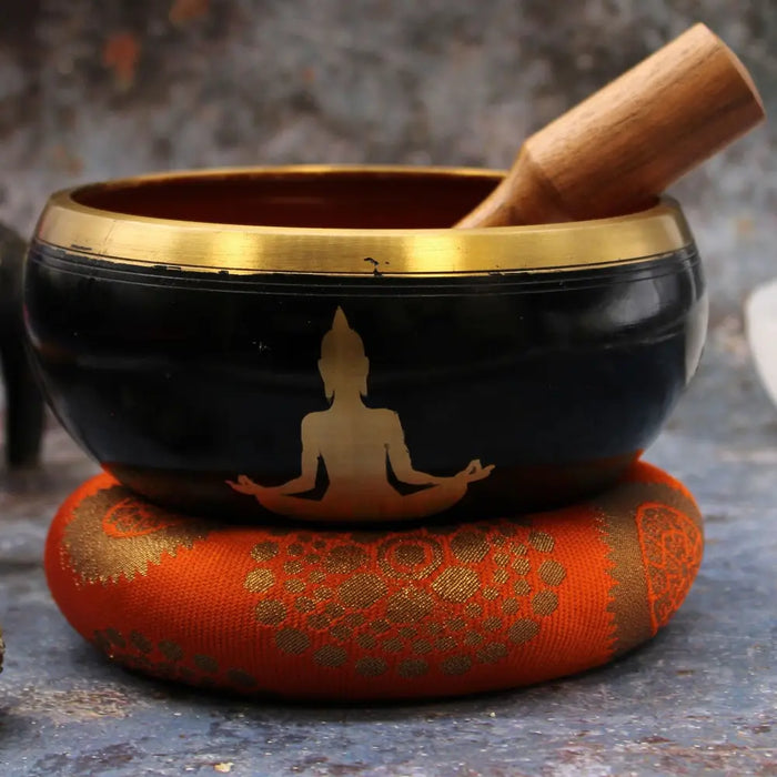 Large black tibetan singing bowl set with gold rim and buddha silhouette resting on orange patterned cushion