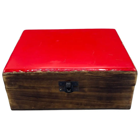 A large ceramic glazed wooden box with a glossy red lid and black metal clasp on a rustic wooden surface