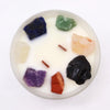A clear glass container with a white candle featuring seven chakras colorful raw gemstones embedded in the wax