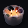 A large white candle with three lit wicks holds colorful raw gemstones representing the seven chakras for harmony