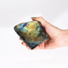 A hand holds a large madagascar labradorite stone 11-14cm with blue green and gold iridescent patterns