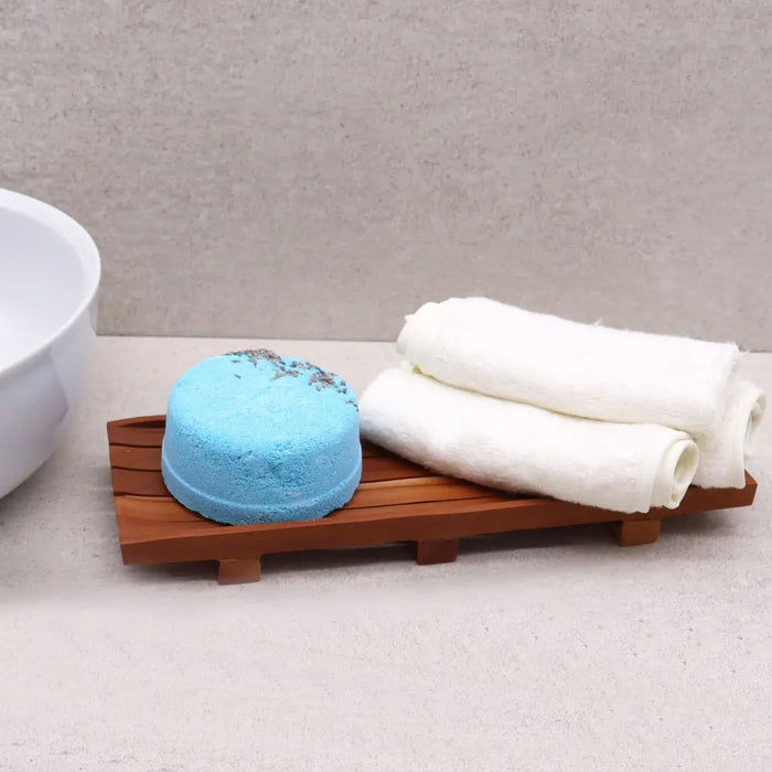 A round blue bath bomb with lavender flakes on a large naseberry wood soap tray with a textured surface