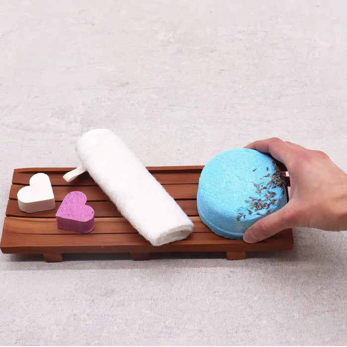 A hand holds a light blue bath bomb with dried lavender on a large naseberry wood soap tray