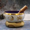 A large yin yang singing bowl set with a white bowl featuring gold floral trim and a purple interior on a patterned cushion