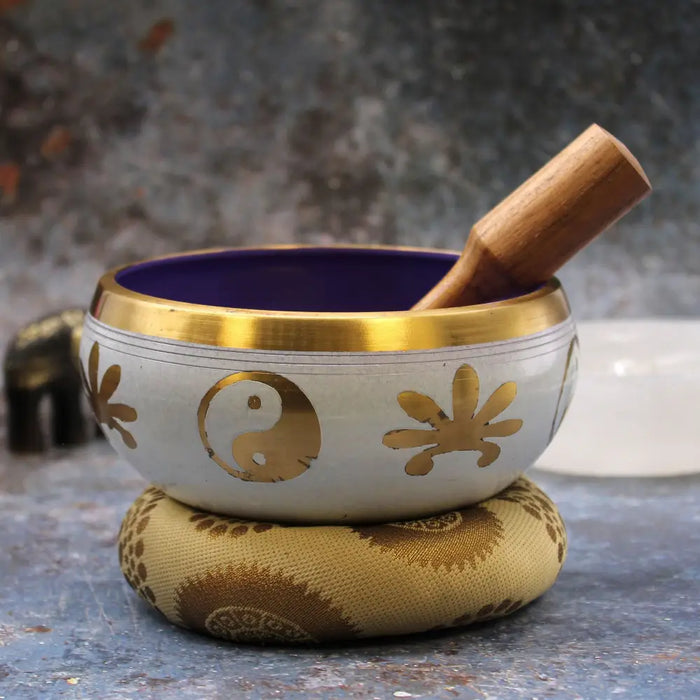 A large yin yang singing bowl set with a white bowl featuring gold floral trim and a purple interior on a patterned cushion