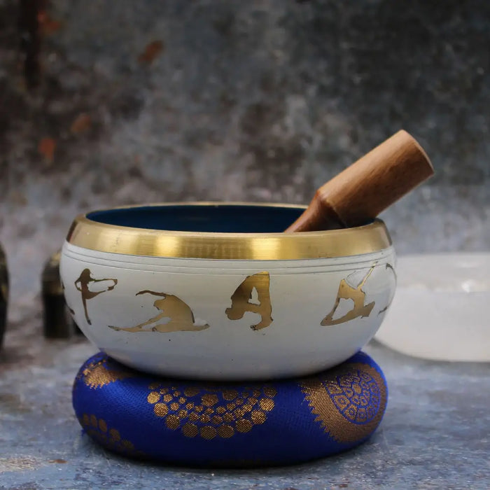 Large white singing bowl with gold trim and engraved figures on blue cushion from yoga moves singing bowl set