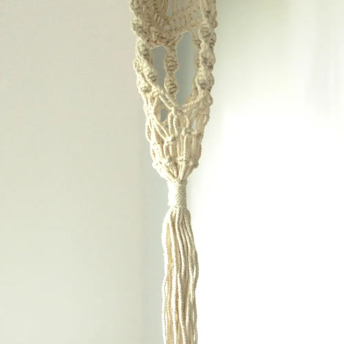 Cream-colored long macrame pot holder with rattan hoop featuring intricate knotting and a tassel at the bottom