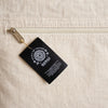 Black rectangular tag with white spirit of travel and nomad logos attached to cream canvas cushion cover by gold zipper pull