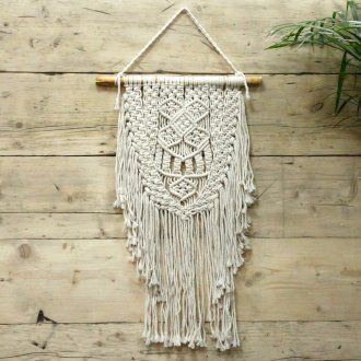 A cream macrame wall hanging with intricate woven patterns and long fringe on a wooden dowel titled abundance