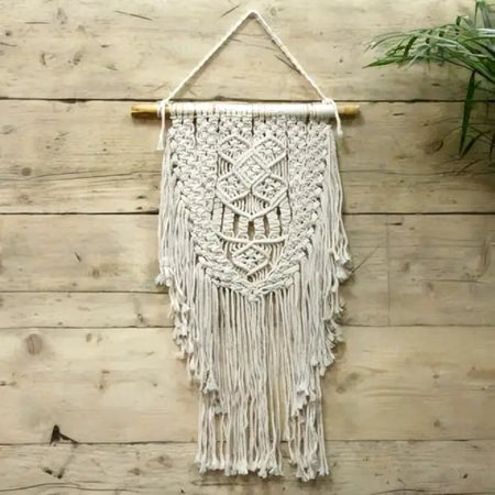 A cream macrame wall hanging with intricate woven patterns and long fringe on a wooden dowel titled abundance
