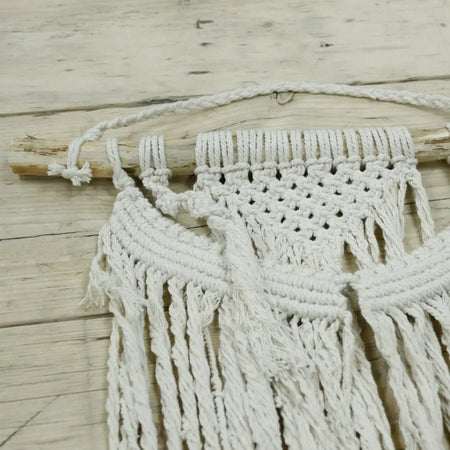 A white macrame wall hanging with woven pattern and long fringe hanging from a wooden dowel called force of nature