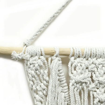 White macrame wall hanging with intricate knotting and tassels suspended from a light wood dowel for weddings
